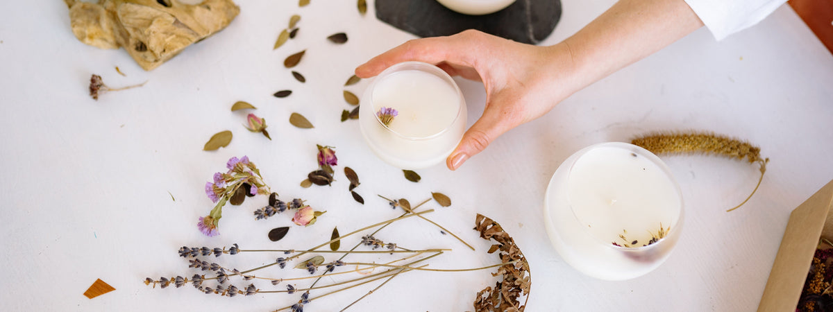 A hand picking up a round white candle in a glass jar surrounded by dried lavender, rosebuds, and botanicals on a white surface, capturing a calm, artisanal vibe.