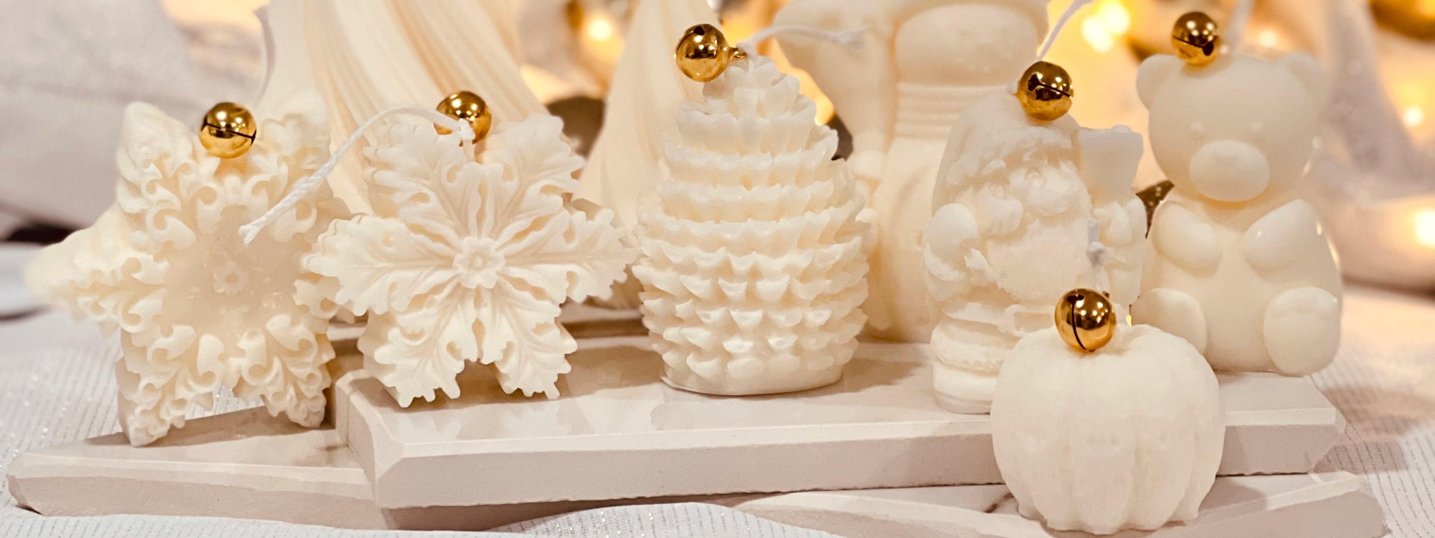 Assorted ivory holiday candles shaped like snowflakes, pine trees, Santa Claus, a bear, and a pumpkin, each topped with a gold ornament bell, styled on neutral tiles with warm ambient lighting.