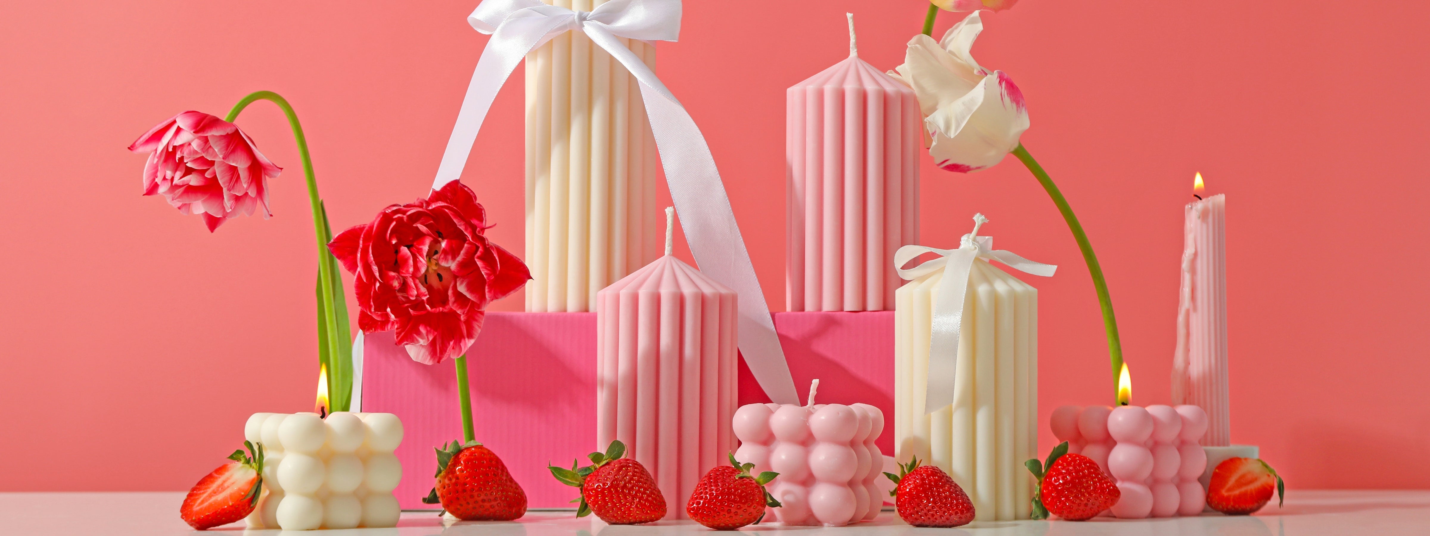 Pink and ivory sculptural candles with strawberries, tulips, and white ribbons arranged against a bright pink background.