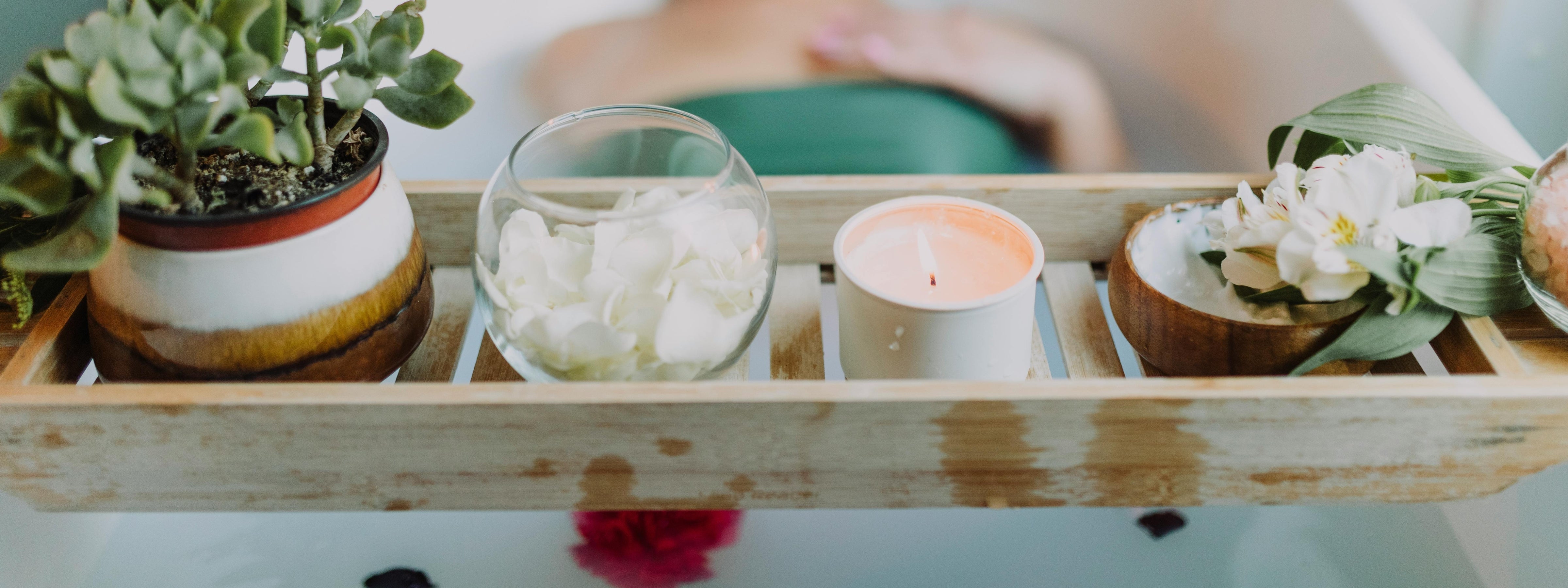 A relaxing bath scene featuring a wooden tray with a potted succulent, a glass bowl of white rose petals, a lit candle in a white jar, and fresh white flowers, with a person soaking in the background.