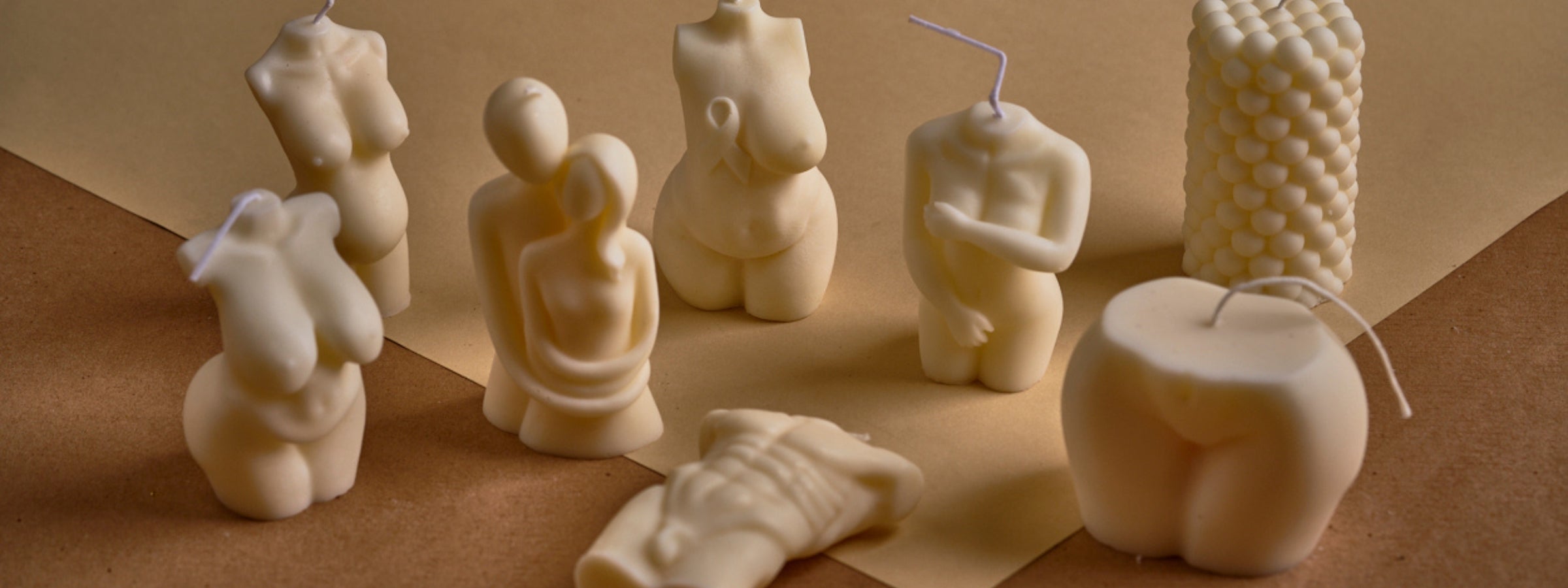 A collection of sculpted cream-colored candles in various artistic shapes, including human torsos, a couple embracing, and geometric designs, displayed on a neutral-toned surface.