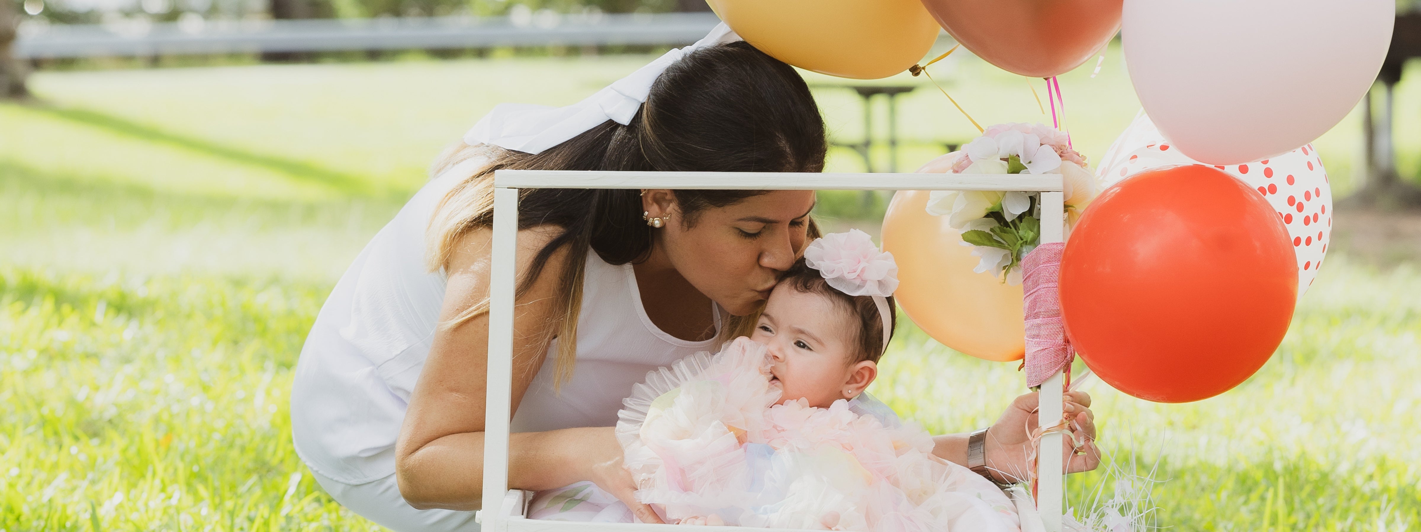 Mother kissing her baby girl during a colorful outdoor celebration with balloons and soft pink floral details.
