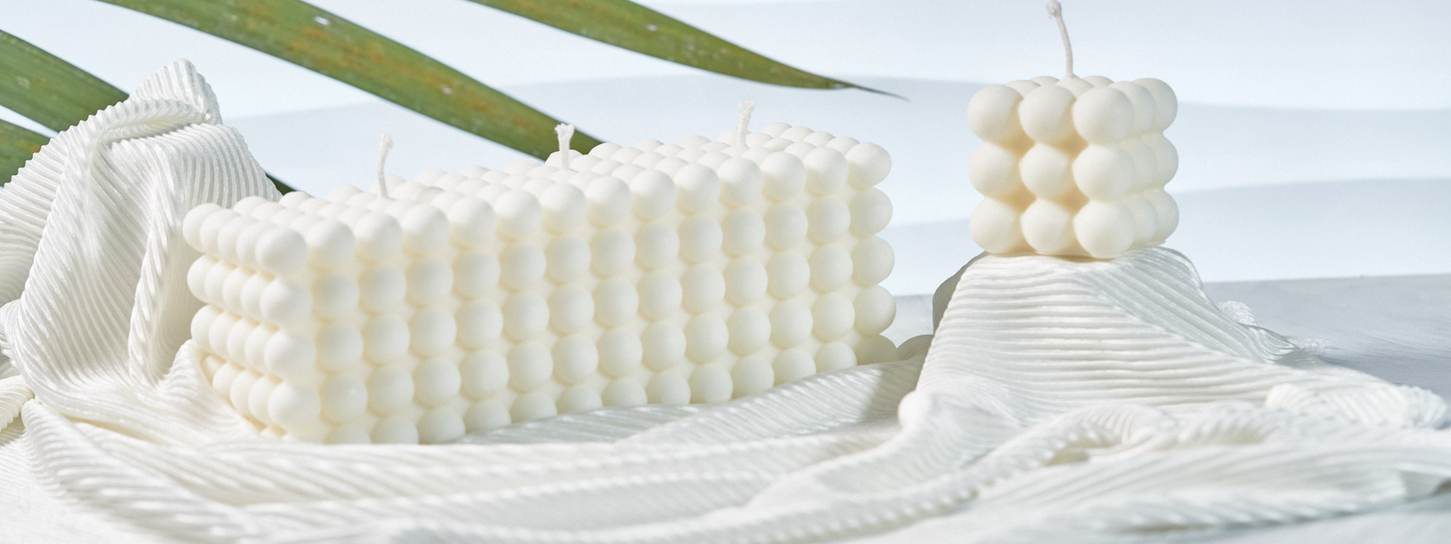 Two ivory bubble candles, one rectangular and one cube-shaped, styled on soft white textured fabric with green leaves in the background for a clean, modern aesthetic.