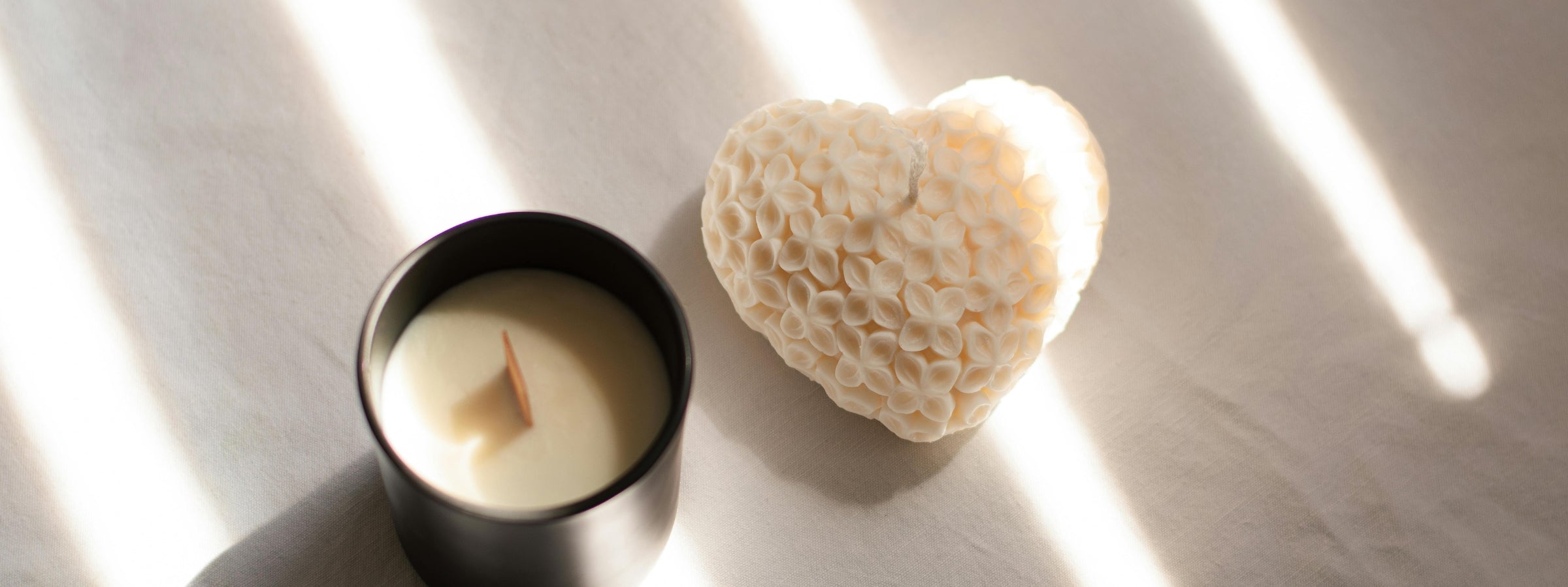 Sunlight dances across a heart-shaped floral candle and a black jar candle, resting quietly on soft fabric.