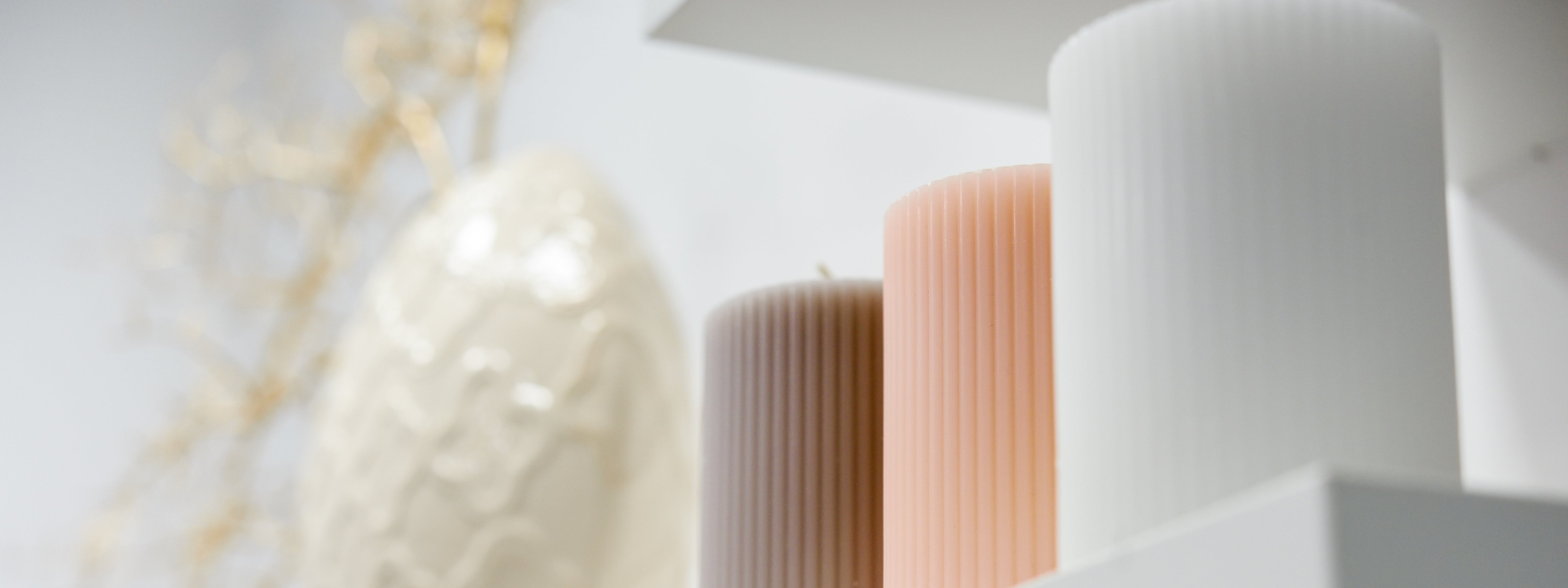 Three ribbed sculptural candles in blush, taupe, and white displayed on a minimalist white shelf.
