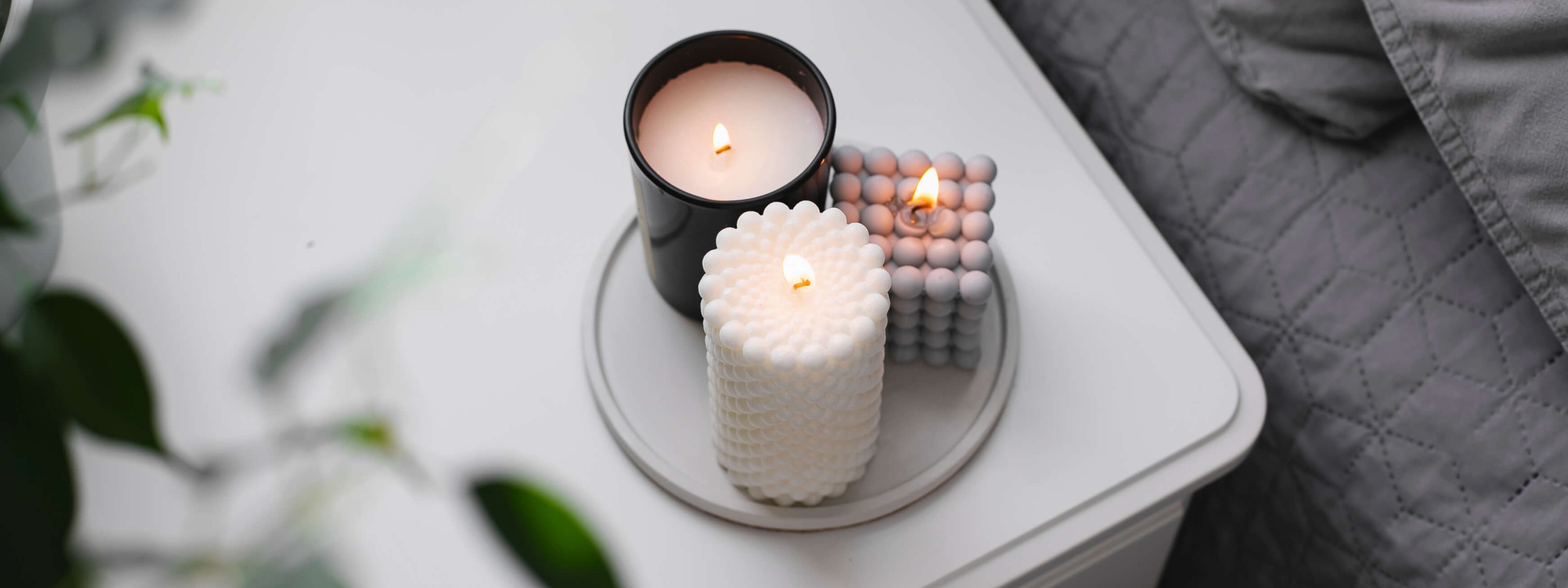 Three lit candles—one in a black glass jar, one white with a beaded texture, and one light gray bubble cube—arranged on a round tray atop a white nightstand beside a quilted gray bedspread.