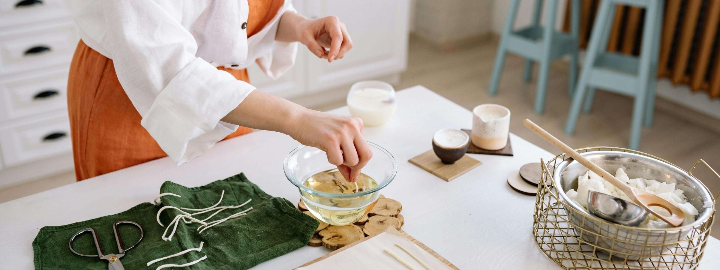 A person is dipping a candle wick into melted wax while preparing handmade candles on a white table with various candle-making tools and ingredients.