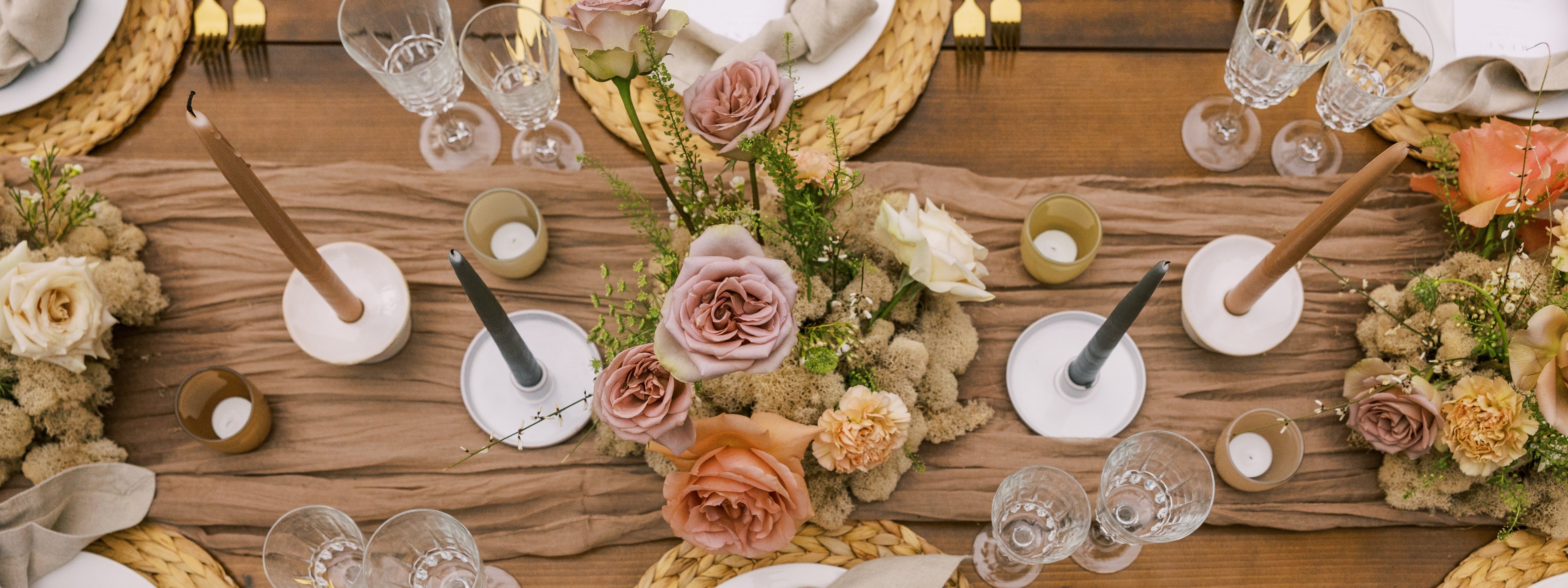 Elegant outdoor table scape featuring taper candles in neutral holders, pastel floral arrangements, woven placemats, and glassware, styled for a seasonal gathering.