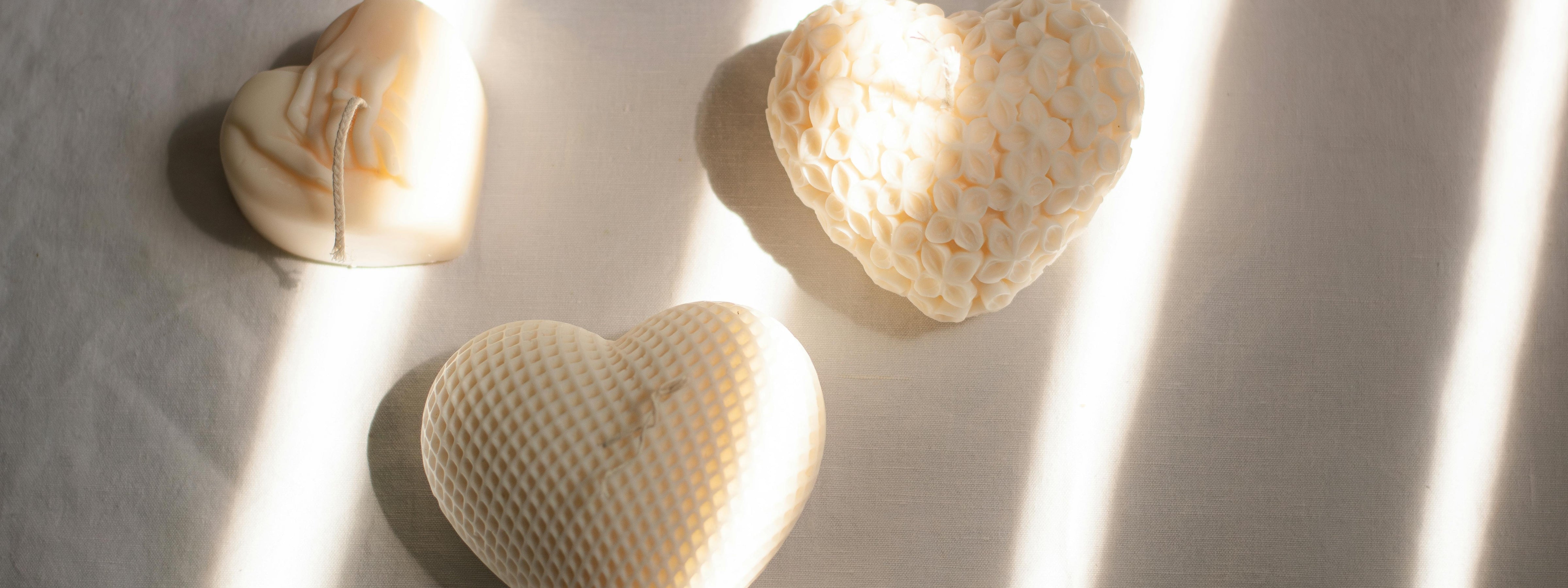 Three heart-shaped ivory candles with unique textures—marbled, floral, and waffle-like—are arranged on a neutral fabric surface, softly lit by striped sunlight.