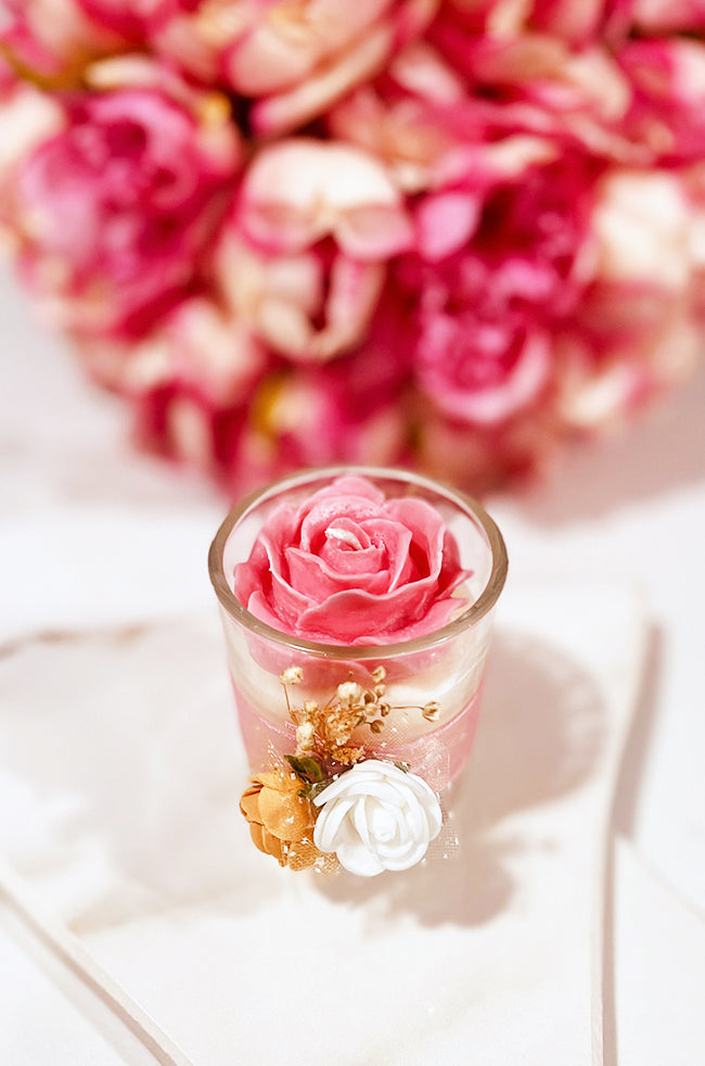 Floating Rose Candle: Pink rose-shaped candle in a glass container, decorated with small white and gold flowers, set against a blurred background of pink roses.