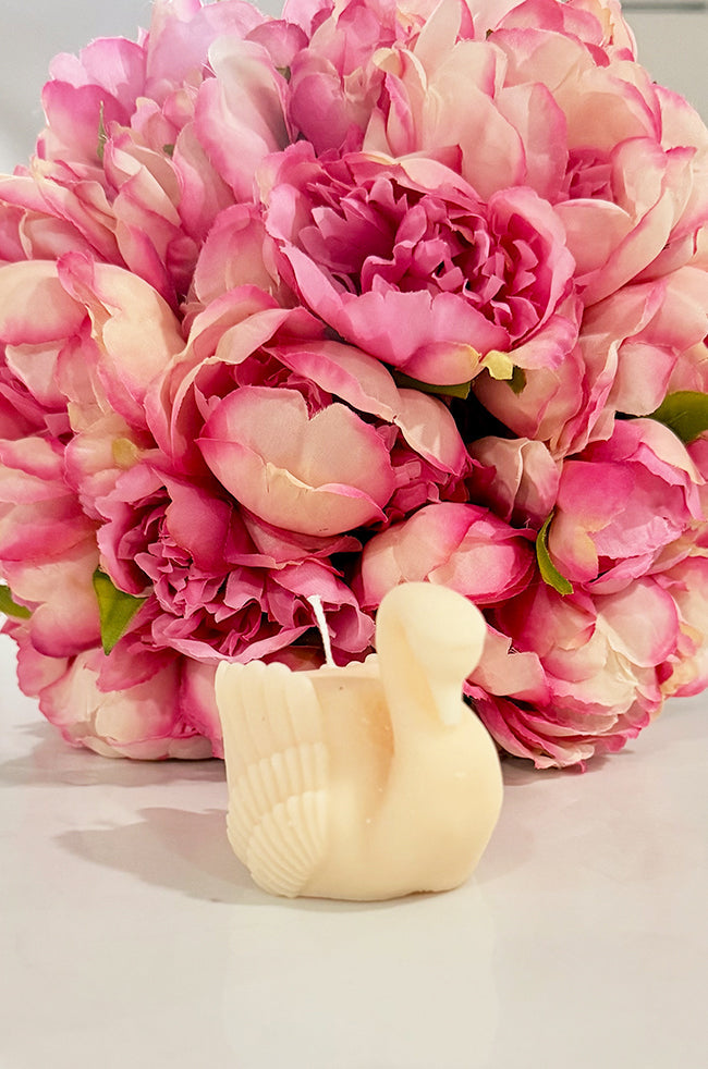 Grace Swan Candle: Handcrafted ivory Grace Swan sculptural candle displayed in front of a bouquet of pink peonies, showcasing its elegant swan silhouette and detailed feather design.
