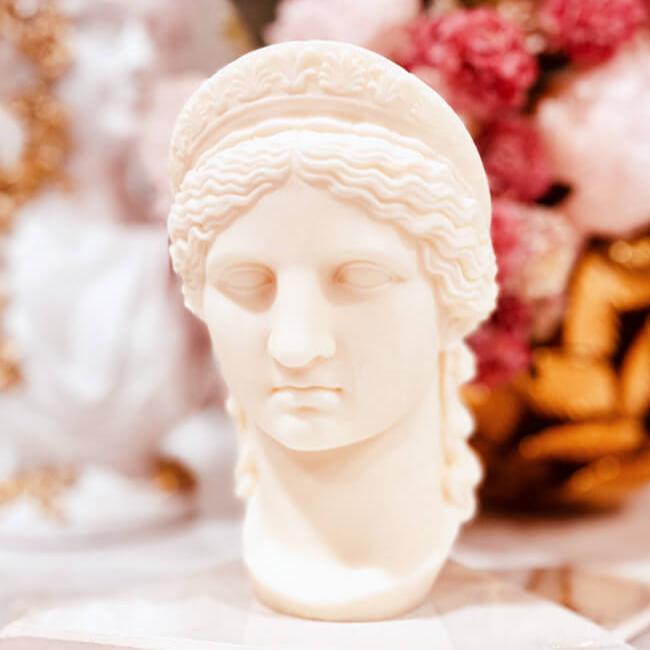 Mother of Olympians Candle: A sculpted candle in the shape of a classical Greco-Roman female bust with intricate hair details and a serene facial expression. The candle is ivory-colored and set against a softly blurred background featuring pink flowers and golden accents.
