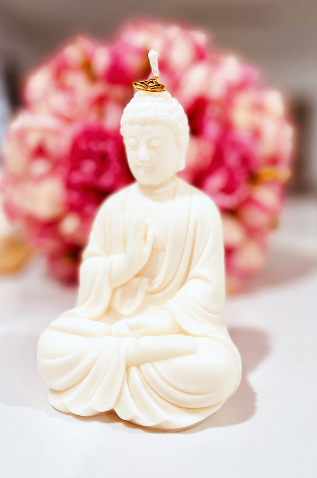 Abhaya Mudra Buddha Candle: A hand-poured candle sculpted in the shape of a seated Buddha in the Abhaya mudra, with a gold lotus charm resting at the base of the wick. The background features soft-focus pink flowers, enhancing the candle's serene and spiritual presence.
