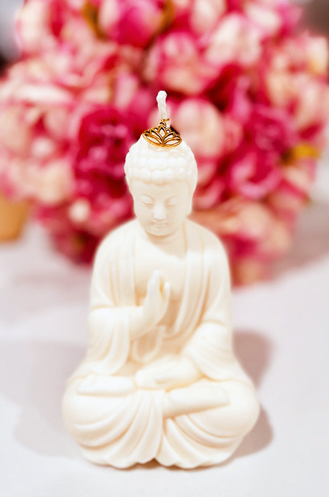 Abhaya Mudra Buddha Candle: A hand-poured candle sculpted in the shape of a seated Buddha in the Abhaya mudra, with a gold lotus charm resting at the base of the wick. The background features soft-focus pink flowers, enhancing the candle's serene and spiritual presence.