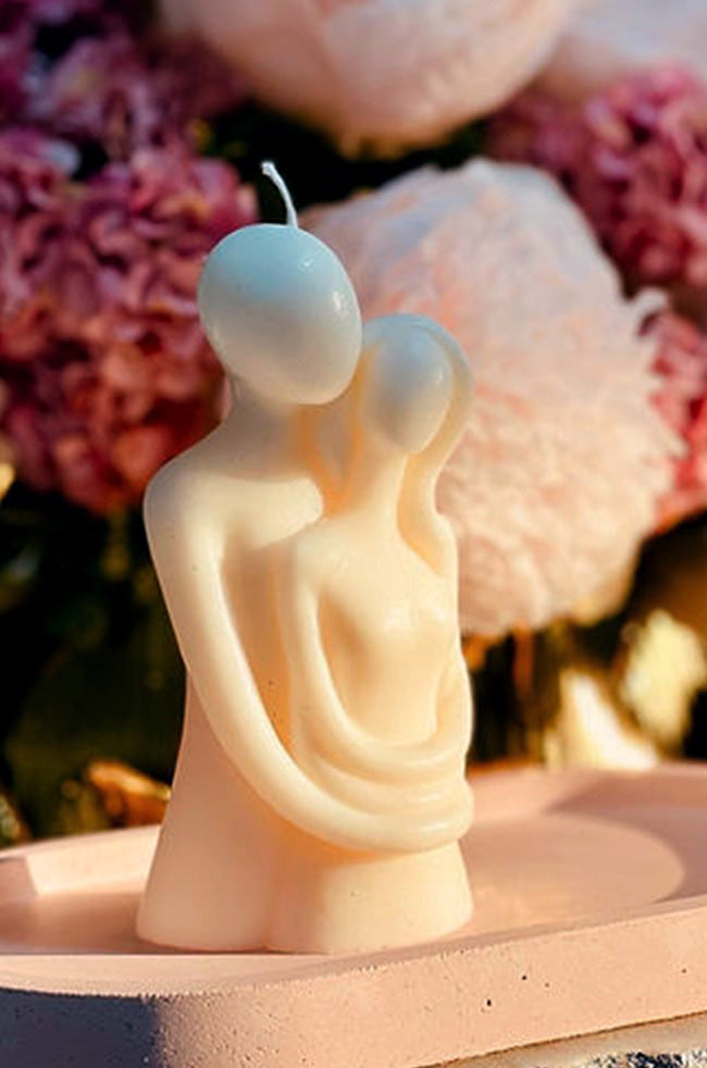 Amore Mio Candle: A sculpted candle depicting an abstract couple embracing, placed on a pink tray with soft pink and purple flowers in the background.