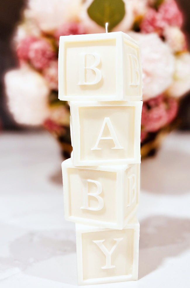 Baby's Building Blocks Candle: A white sculpted candle that looks like stacked baby blocks spelling out "BABY," set against a soft background of pink and white flowers.