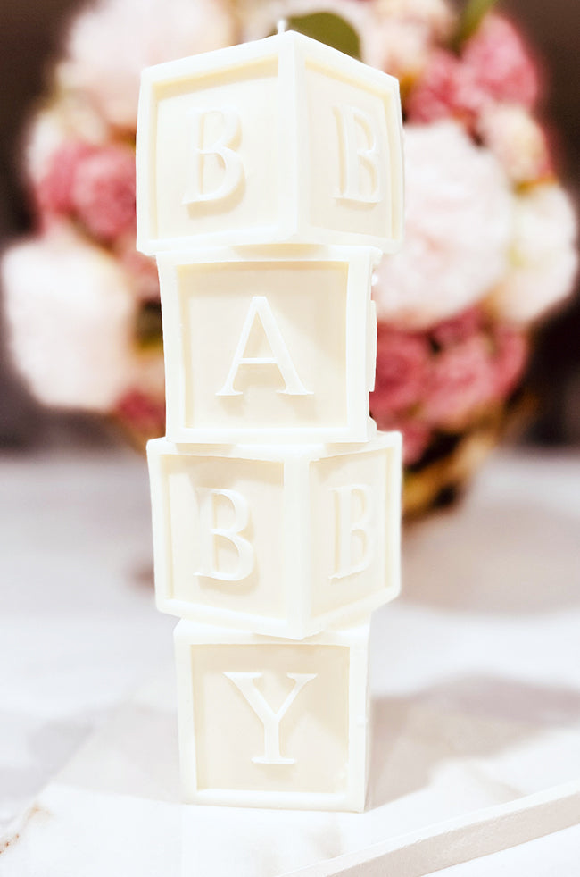 Baby's Building Blocks Candle: A white sculpted candle that looks like stacked baby blocks spelling out "BABY," set against a soft background of pink and white flowers.