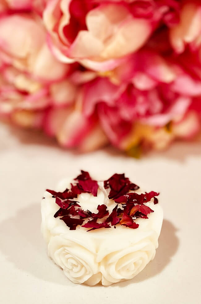 Bed Of Roses Candle with Rose Petals: A white rose-shaped candle topped with dried red rose petals, set against a soft pink floral background, evoking a romantic and elegant atmosphere.