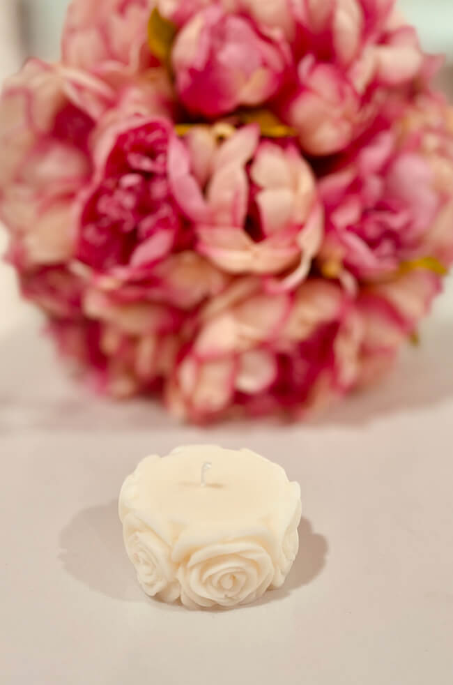 Bed Of Roses Candle Plain: A white rose-shaped candle with intricate floral detailing, placed on a light surface with a blurred bouquet of pink flowers in the background.