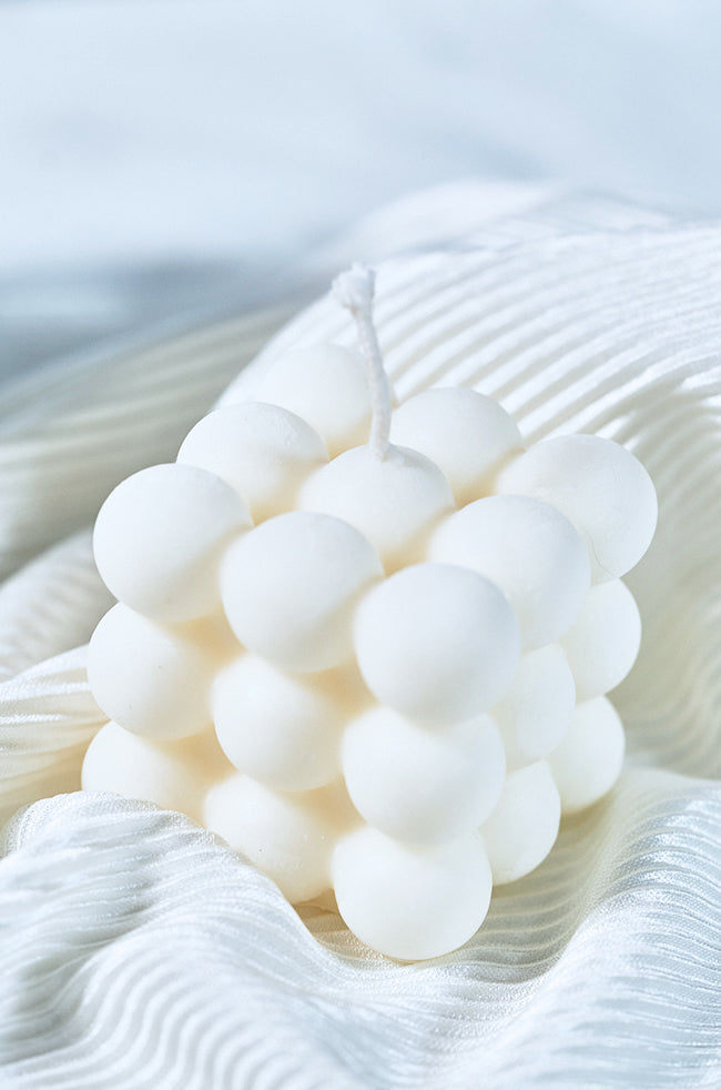 Bubble Candle Plain: A white bubble cube candle with a smooth, rounded surface pattern, placed on soft, white textured fabric under natural light.