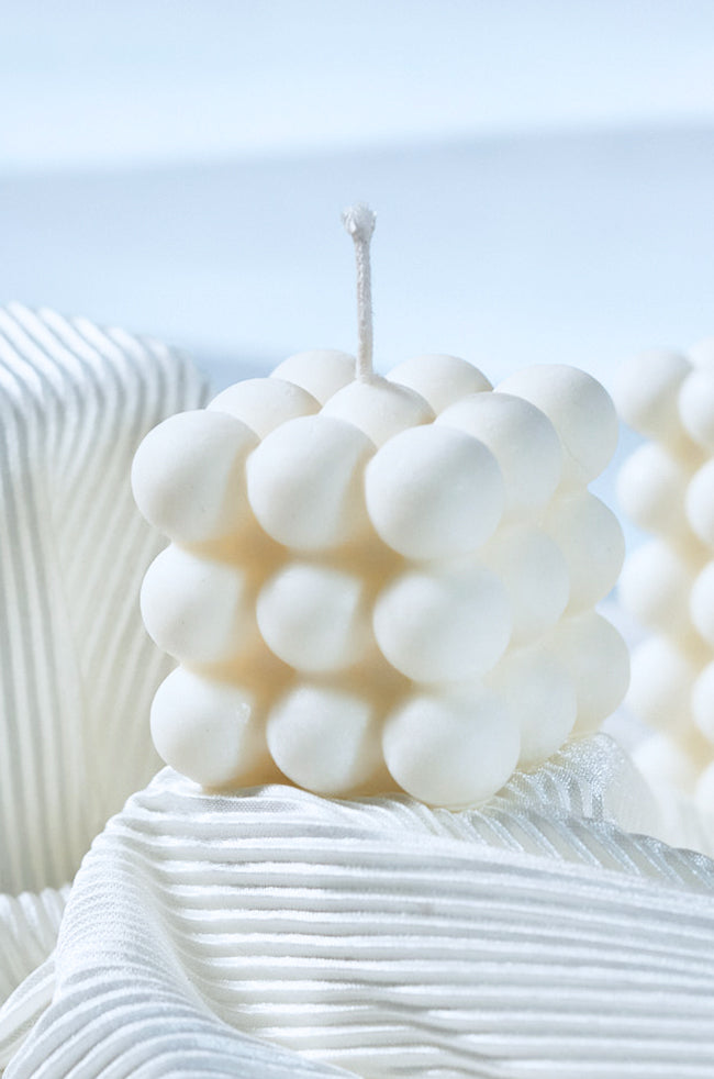 Bubble Candle Plain: A white bubble cube candle with a smooth, rounded surface pattern, placed on soft, white textured fabric under natural light.