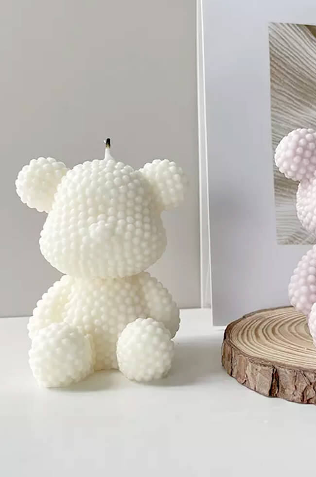 Bubbly Bear Candle: A white bear-shaped candle with a textured, bubble-like surface, placed on a white surface next to a wood slice and a decorative photo frame.