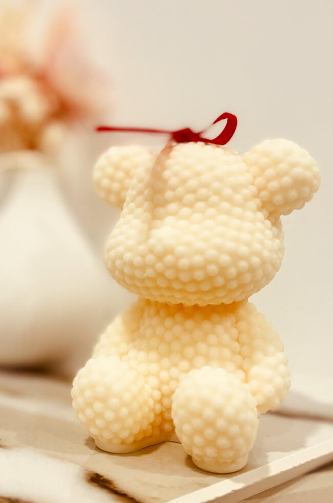 Bubbly Bear Candle: A cream-colored bear-shaped candle with a bubbly texture, adorned with a small red ribbon on top, sitting on a marble surface.