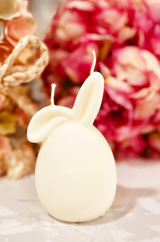 Chilling Bunny Candle: A white sculpted candle shaped like a bunny with long ears, placed on a light surface with soft pink and peach flowers in the background.