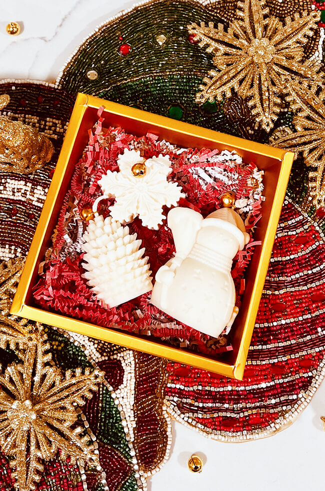 Christmas Gift Box Candles: A festive gift box with gold edges containing three white sculpted candles shaped like a snowflake, a pine tree, and a snowman. The candles are in red and white crinkle paper with small gold bells. The box is set on a decorative, beaded holiday table runner featuring gold, green, and red patterns.