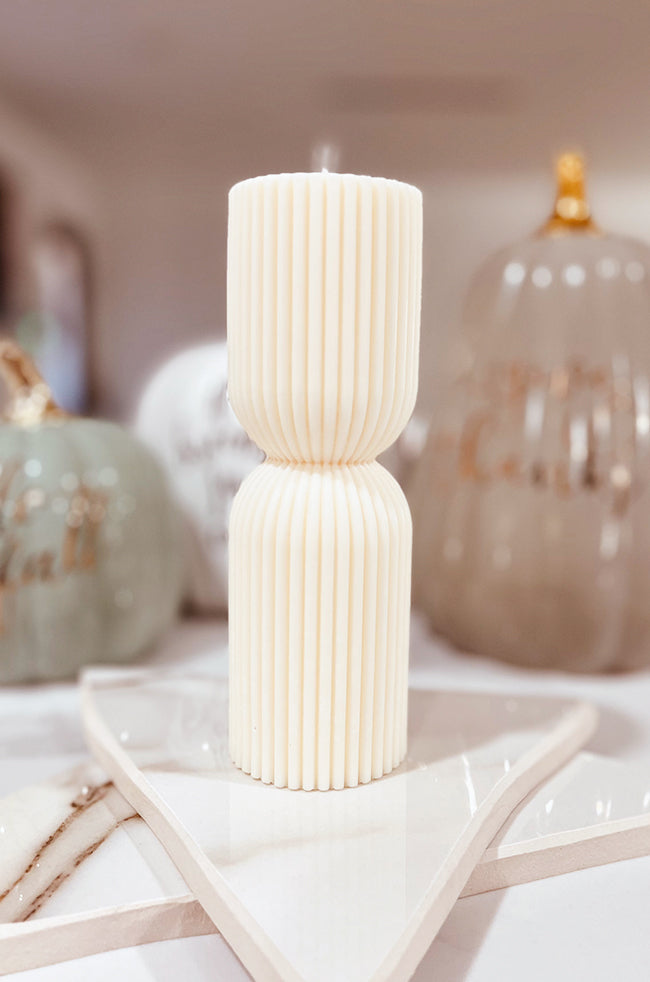 Colliding Column Candle Medium: A tall, sculpted white candle with vertical ridges and a symmetrical hourglass shape, displayed on a star-shaped tray with fall decor in the background.