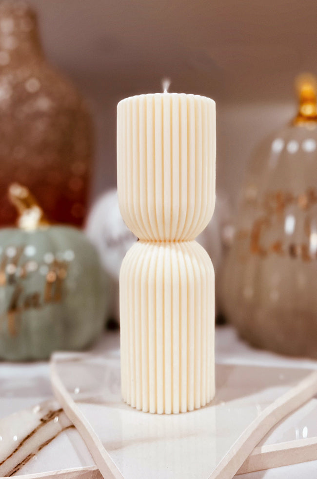 Colliding Column Candle Medium: A tall, sculpted white candle with vertical ridges and a symmetrical hourglass shape, displayed on a star-shaped tray with fall decor in the background.