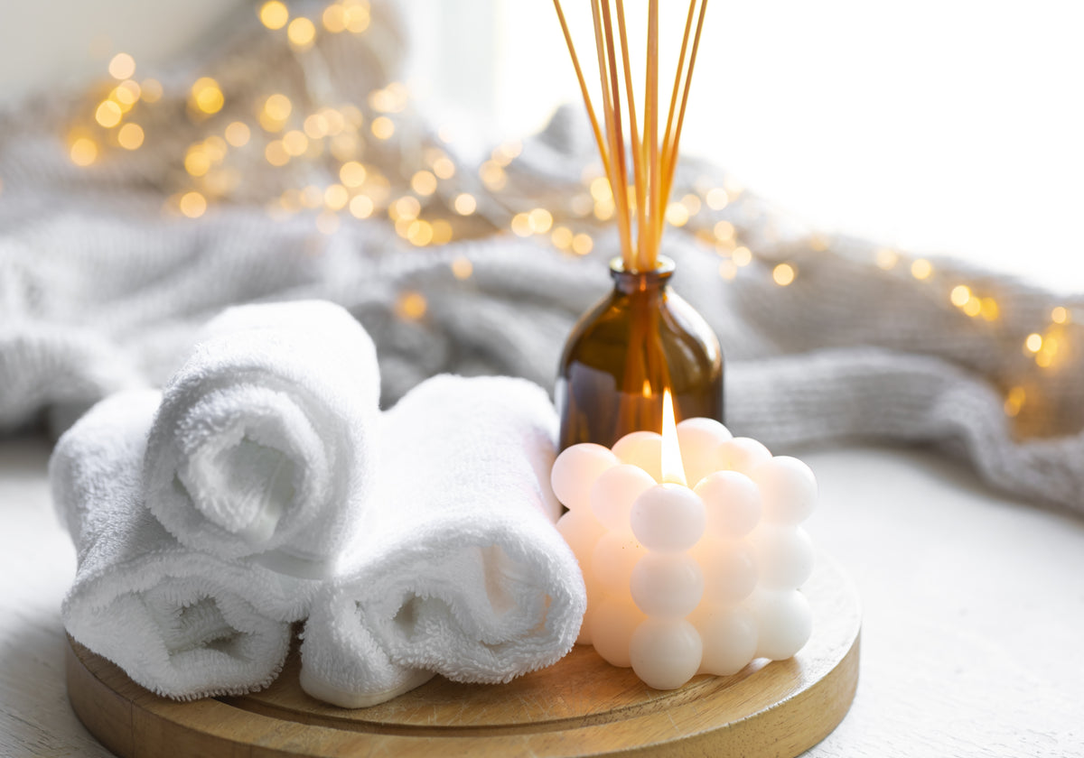 A spa-style still life with three rolled white towels on a round wooden tray beside a lit white bubble candle and an amber glass reed diffuser bottle, set against a soft gray knit blanket and warm golden bokeh lights.