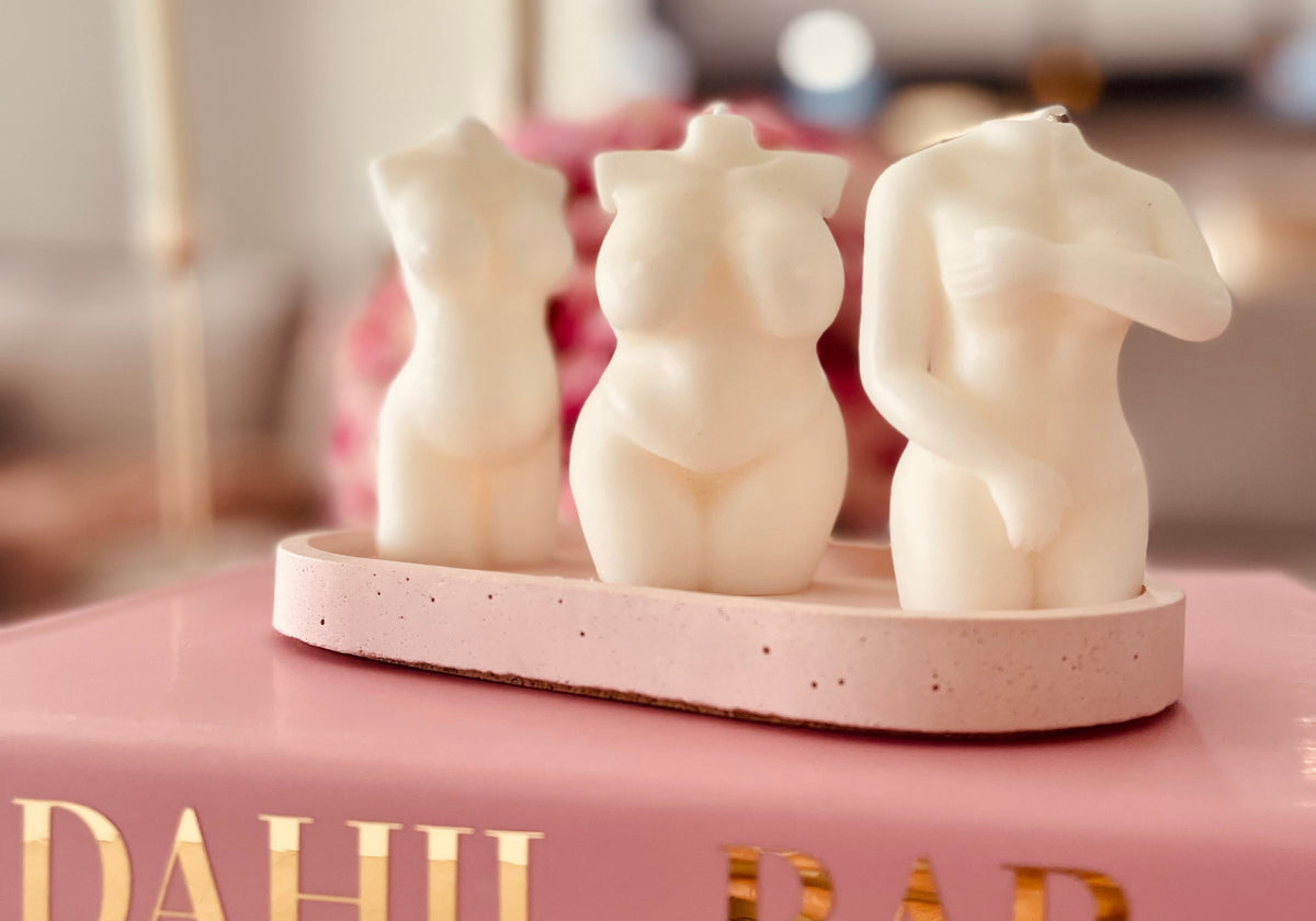 Three ivory female-form sculptural candles displayed on a pink tray over stacked Dahil Bar books.