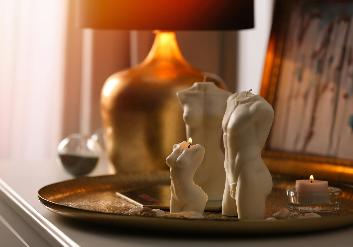 Two cream-colored male and female torso candle sculptures on a gold tray, styled on a table next to a lit gold lamp and a small tea light candle.
