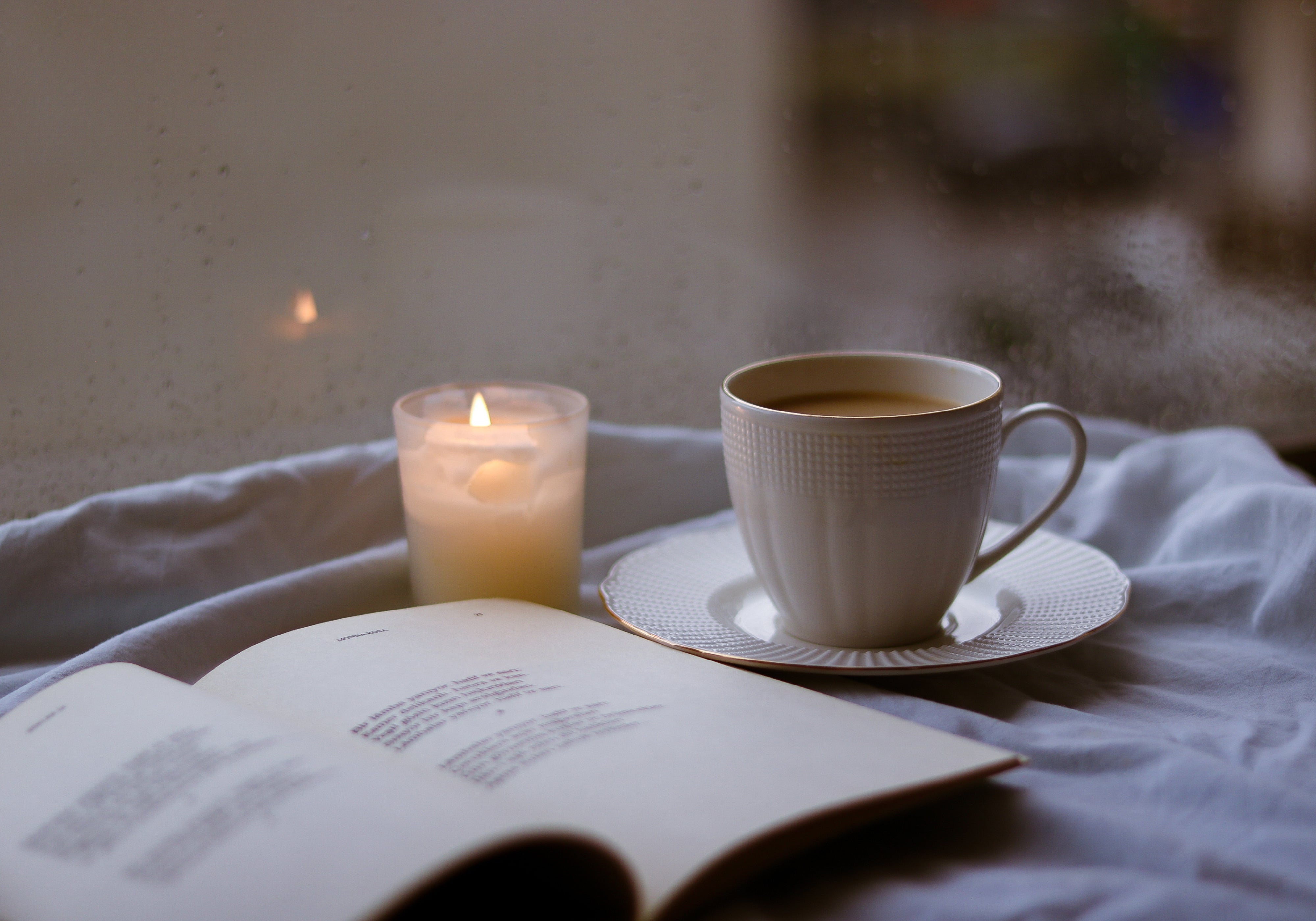 Lit candle beside a warm cup of tea and an open book on soft bedding, creating a calm, cozy fall atmosphere.