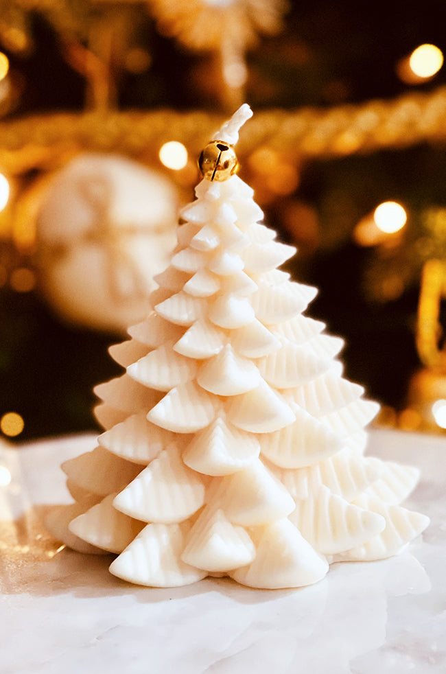 Fiery Tree Candle Large: Ivory sculptural candle shaped like a Christmas tree, topped with a small gold bell, styled in front of festive holiday lights.