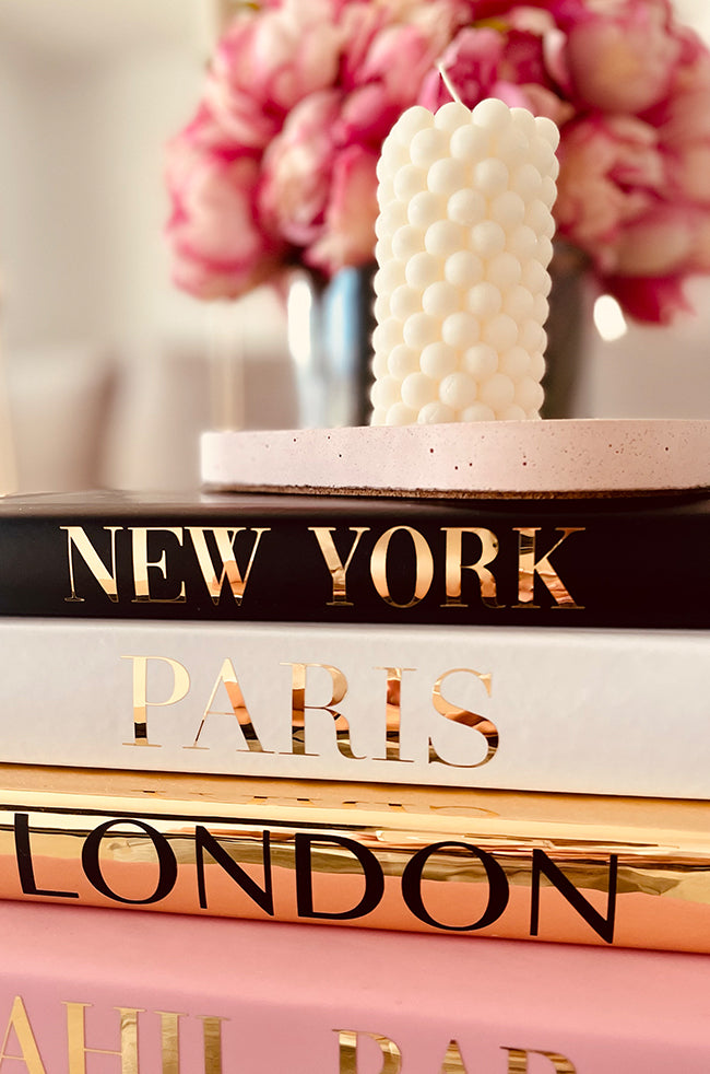 Fire Beads Candle: Ivory stacked bead pillar candle on a pink tray, displayed atop a stack of books labeled New York, Paris, and London, with pink flowers in the background.