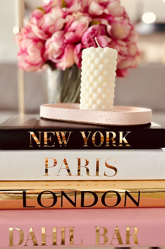 Fire Beads Candle: Ivory stacked bead pillar candle on a pink tray, displayed atop books labeled New York, Paris, London, and Dahil Bar, with pink flowers in the background.