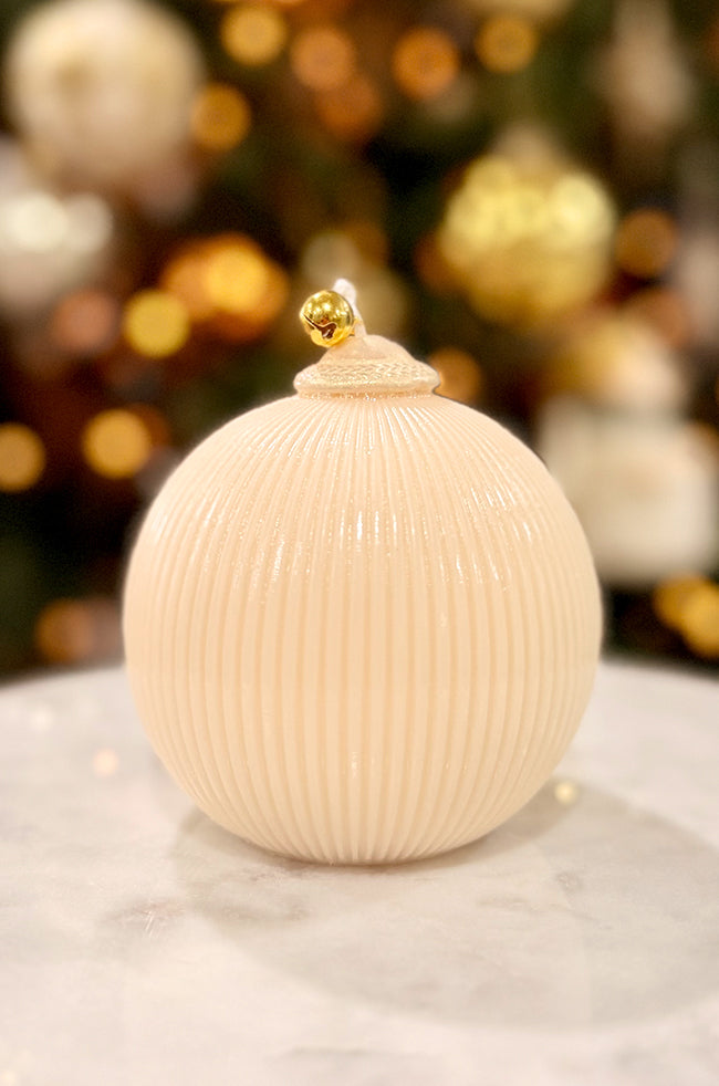 Golden Celestial Ornament Pear Candle: A pearl-white ribbed ornament candle topped with a small gold bell, displayed on a marble surface with warm golden Christmas lights in the background.