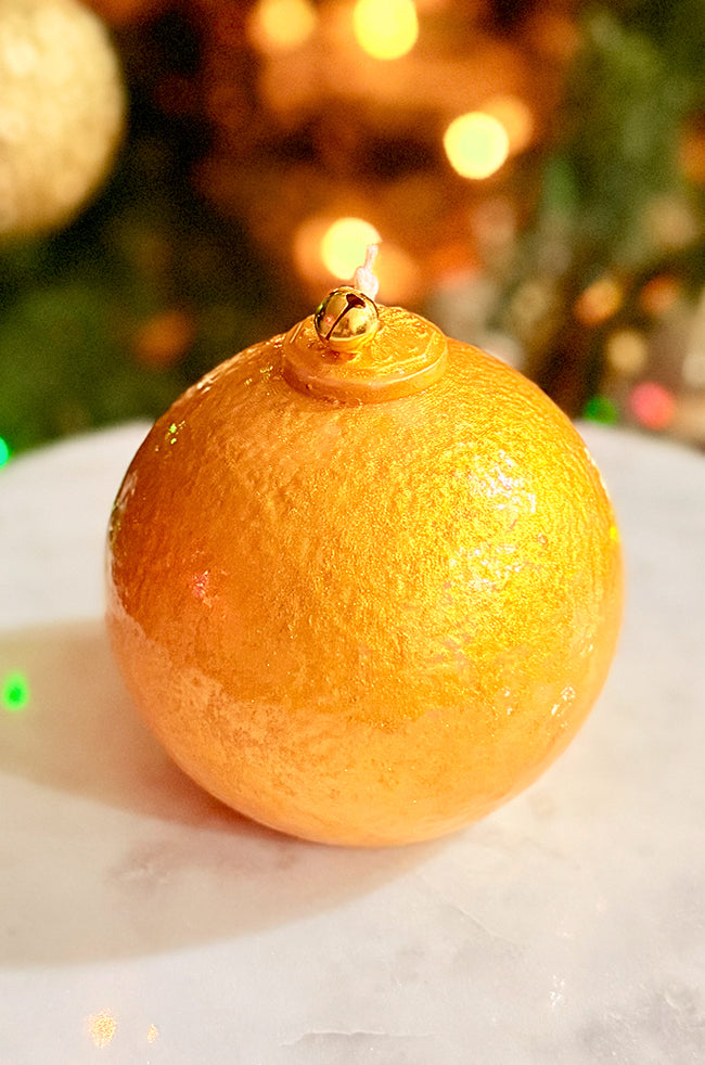 Golden Luxe Ornament Candle: A hand-painted gold ornament candle with a small gold bell on top, displayed on a white marble surface with warm holiday lights in the background.