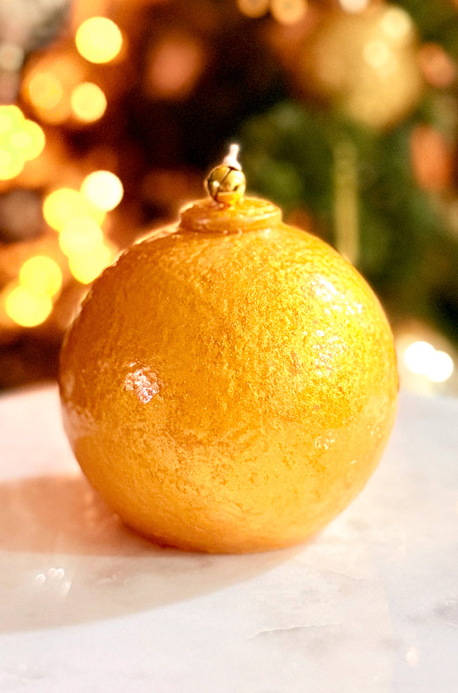 Golden Luxe Ornament Candle: Close-up of a metallic gold ornament candle featuring a small gold bell at the wick, shown against soft, glowing Christmas tree lights.
