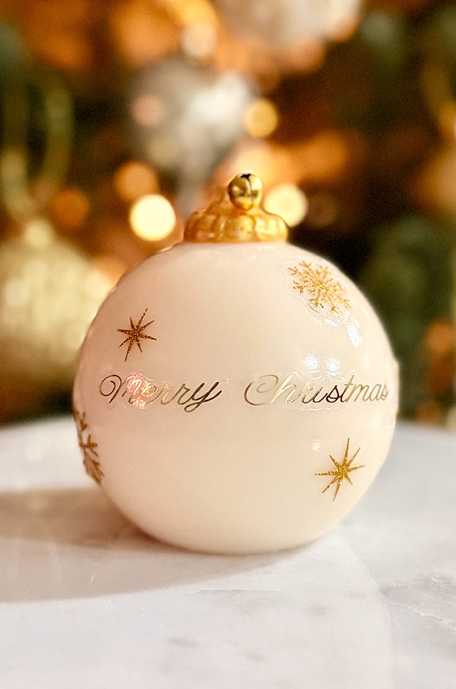 Golden Merry Ornament Candle: Ivory ornament candle with gold hand-painted stars and a “Merry Christmas” inscription, topped with a golden bell, styled on a marble surface with warm holiday lights in the background.
