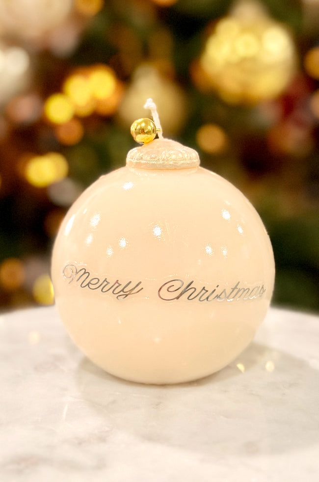 Golden Merry Ornament Candle: Ivory Christmas ornament candle with glossy finish, gold bell topper, and “Merry Christmas” lettering, displayed on marble with soft holiday lights.