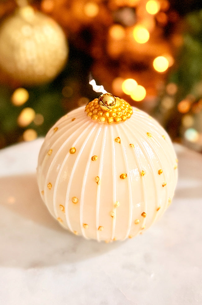 Golden Royale Ornament Candle: Golden Royale Ornament candle featuring ivory ribbed detailing and scattered gold droplets, topped with a gold bell, styled in front of blurred golden Christmas décor.