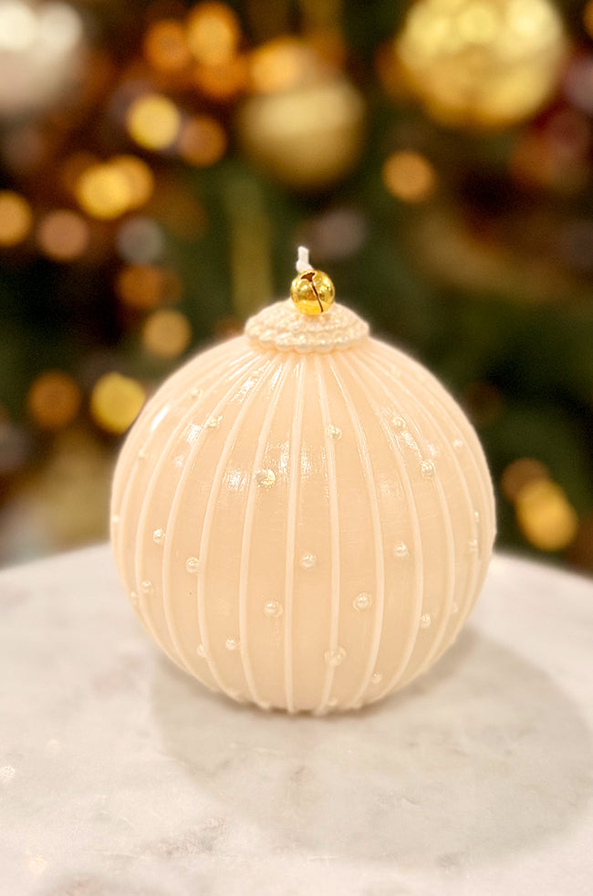 Golden Royale Ornament Candle: Ivory ornament-shaped candle with vertical ridges and pearl-like painted accents, topped with a small golden bell, displayed on a marble surface with warm holiday lights in the background.