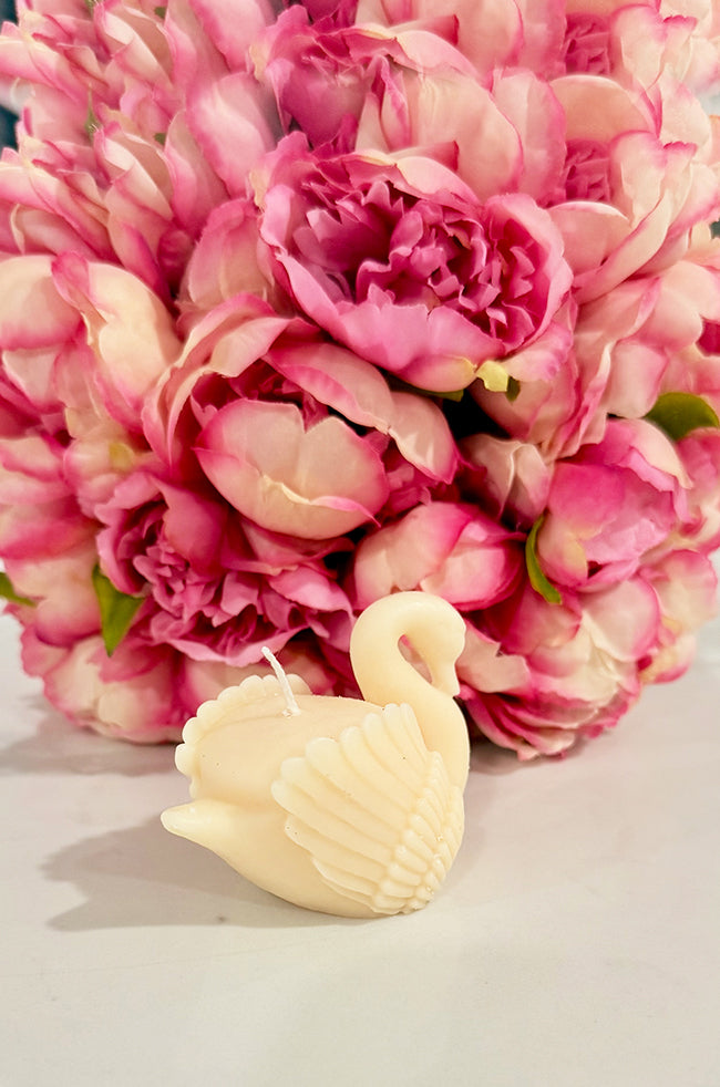 Grace Swan Candle: Side view of the handcrafted ivory Grace Swan candle highlighting its intricate feather details with soft pink peonies blurred in the background.