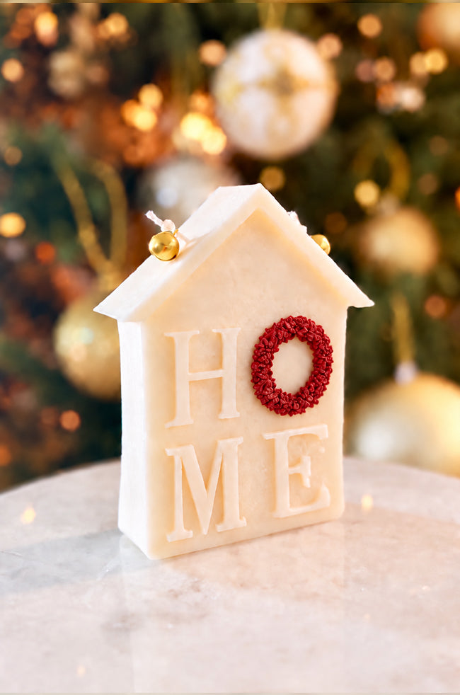 Home for Christmas Candle: Angled view of the Home for Christmas sculptural candle featuring HOME lettering, with a red wreath detail and two golden bells against festive holiday décor.