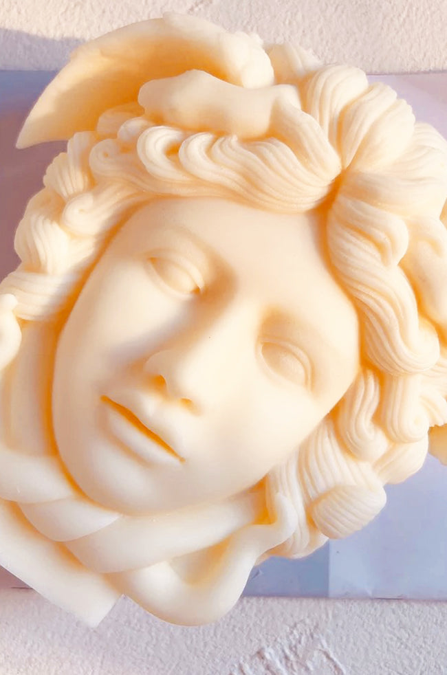 Medusa Candle: Overhead view of the Medusa candle laid flat on a neutral surface, highlighting the detailed snake carvings in the hair and symmetrical face structure.
