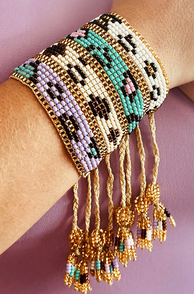 Orchid: A wide, handwoven bracelet featuring pastel and gold seed beads arranged in geometric and animal-inspired patterns, with braided golden cords and beaded tassels hanging from the ends.