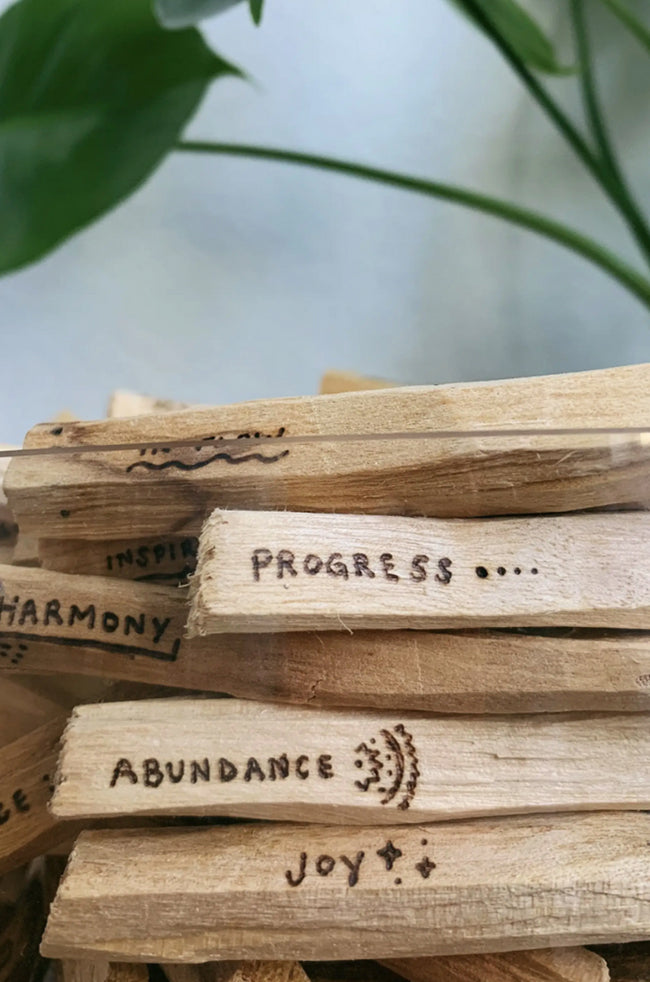 Palo Santo: Stack of Palo Santo sticks engraved with uplifting words like “Abundance,” “Joy,” and “Progress,” styled beside greenery.